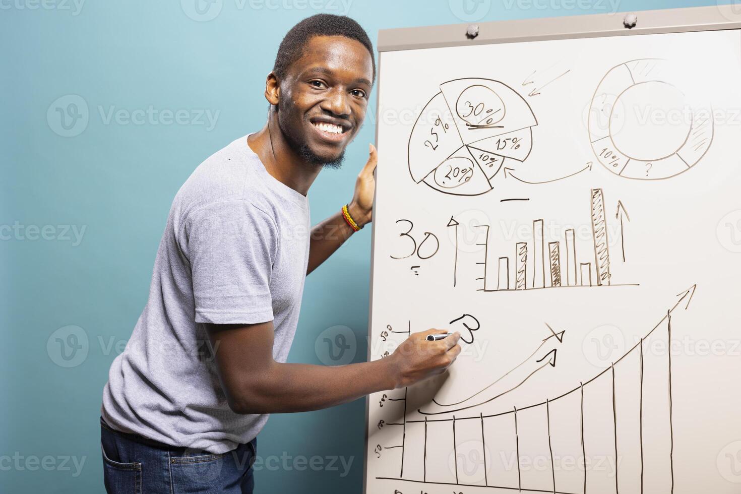 Young black man writing on whiteboard and explaining a chart, standing against a blue studio background. Smiling african american guy holding a pen and using a marker board to showcase a graph. photo