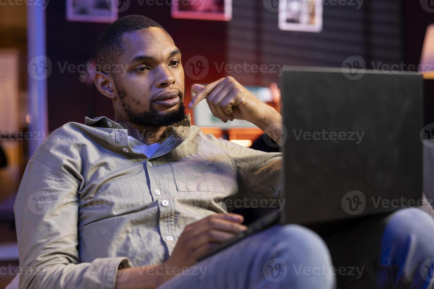 African american adult working remotely from his apartment on laptop, focusing on tasks completion while he lays on a bean bag. Blending productivity and relaxation, browsing the web. photo