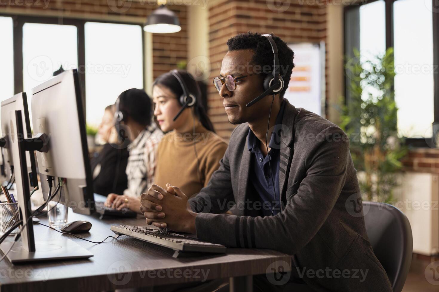 Serious male office worker concentrating on business analytics on his computer in brick wall office. Focused black businessman with clasped hands, wearing a headset, and looking at his desktop monitor. photo