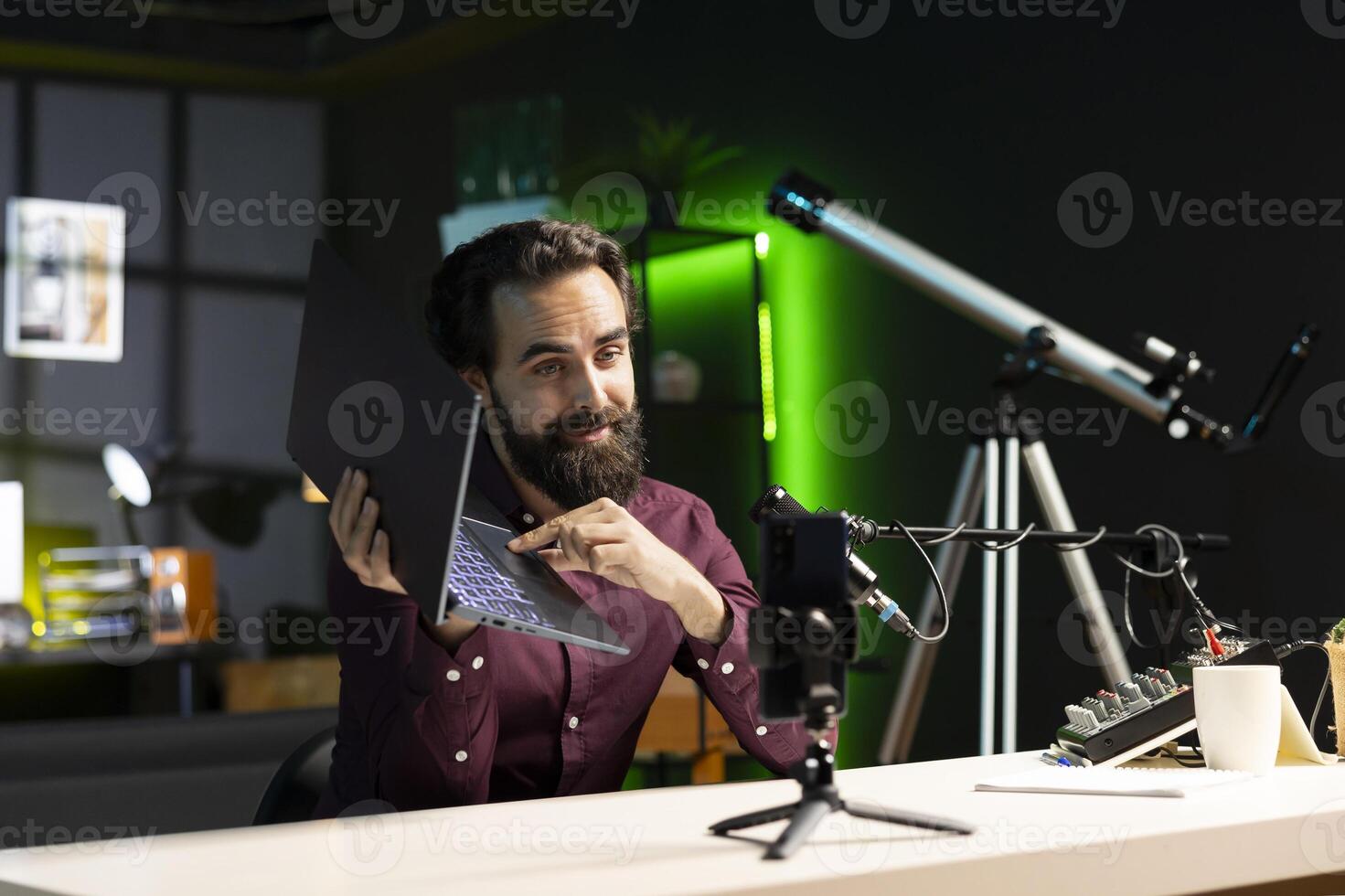 Media star in studio endorsing laptop from brand sponsoring , filming with smartphone on tripod. Tech expert does influencer marketing by promoting notebook, recording with phone photo