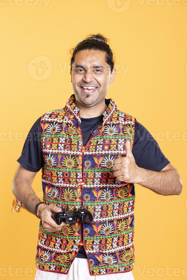 Portrait of overjoyed indian gamer showing thumbs up sign, holding controller, playing videogames. Ecstatic person with gamepad delighted to do gaming, doing approving hand gesture, studio backdrop photo