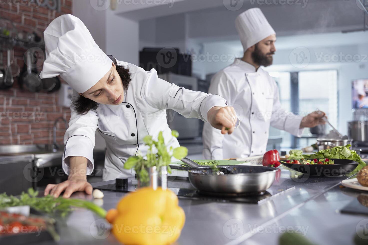 In a kitchen, woman chef perfecting a gourmet meal by adding fresh herbs, balancing flavors with basil and other condiments. Kitchen counter stocked with vegetables and food prep essentials. photo