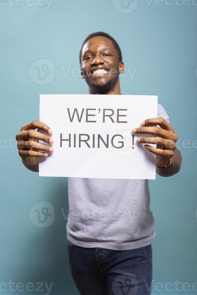 Cheerful male recruiter presenting a hiring sign in a studio, looking for new talent to fill business positions. Excited black man holding a paper towards camera, promoting job vacancies. photo
