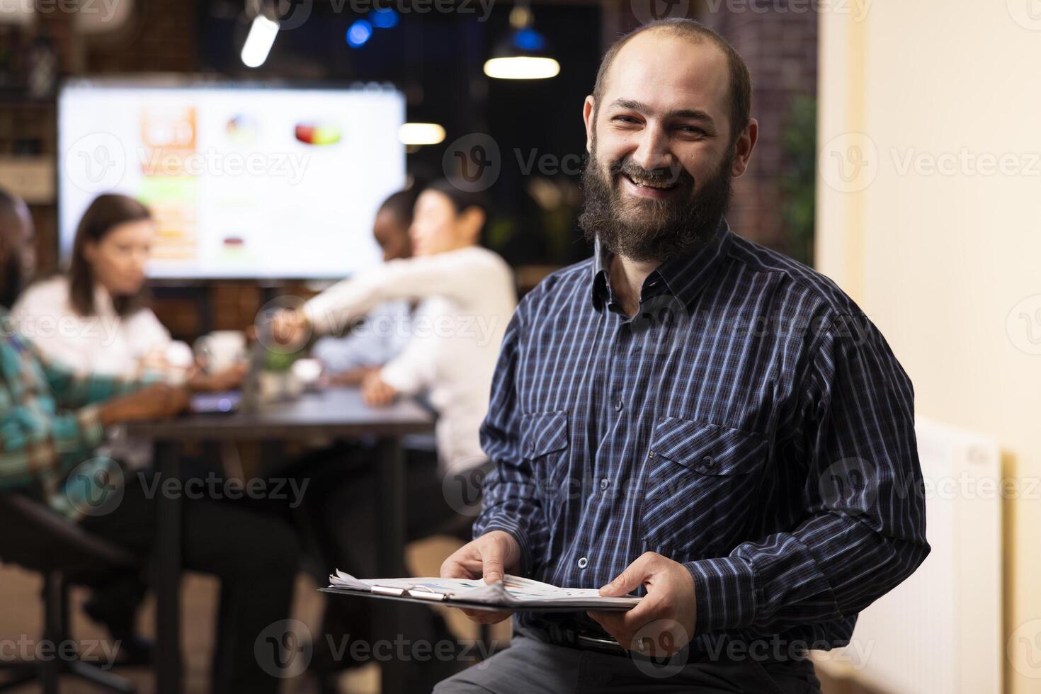 Portrait of caucasian male executive with clipboard, sitting in office, overseeing startup team. The evening setting, brick wall and digital devices emphasize a productive, modern workspace. photo