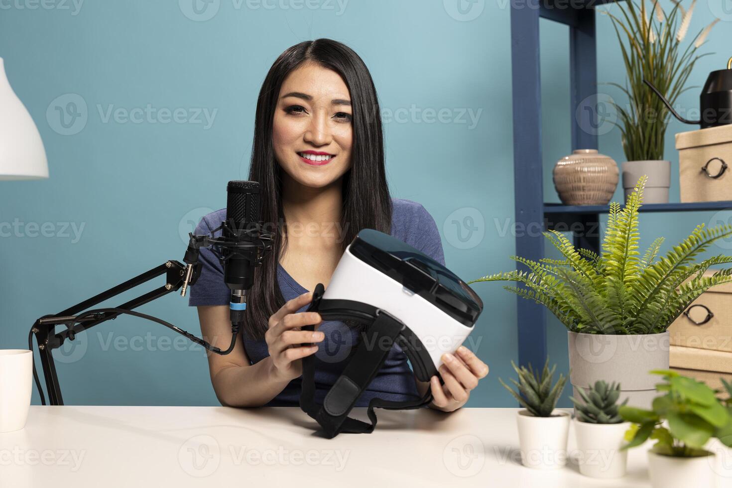 Asian woman vlogger seated at her desk in a studio, holding a VR headset while speaking into a microphone. She is presenting the virtual reality product, reviewing it for her online audience. photo