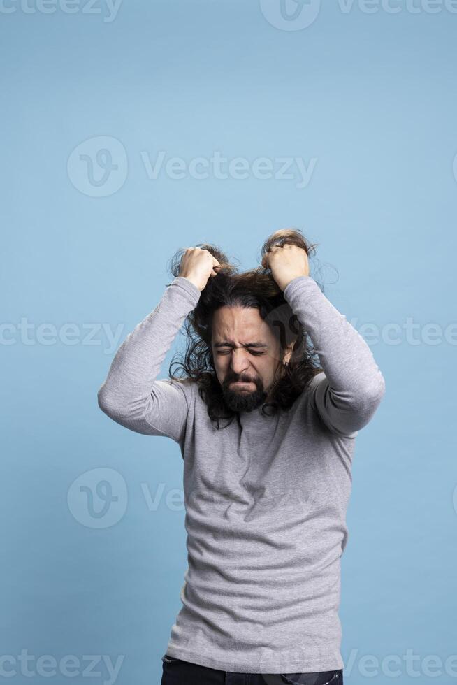Distraught upset guy acting crazy and pulling his hair in front of camera, being overwhelmed and furious with intense emotions. Annoyed man with an aggressive reaction and rage in studio. photo