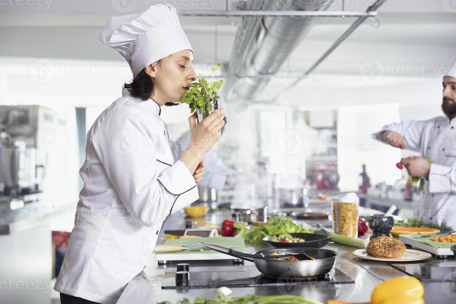 Dedicated female chef smelling the aroma of herbs and greenery, using tasty fresh rosemary or parsley leaves and sprinkling in the pan. Culinary expert cooking dishes with raw materials, gastronomy. photo