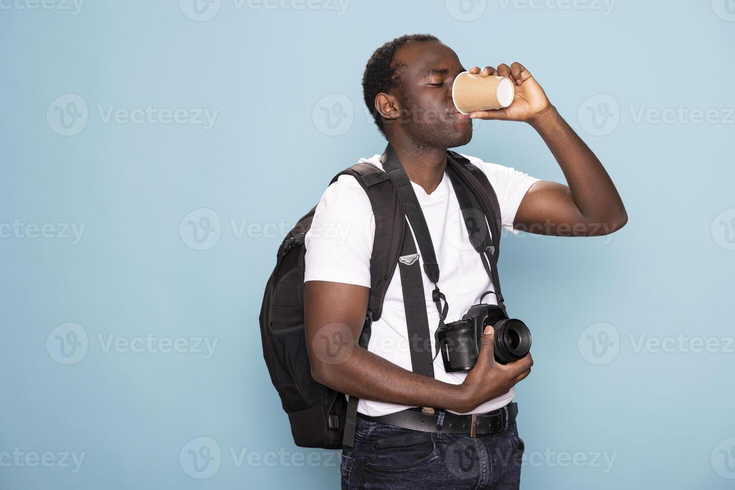 Black photographer in casual clothing carrying backpack and enjoying a hot beverage. African american man with a camera around his neck takes a sip of coffee while standing against isolated background. photo