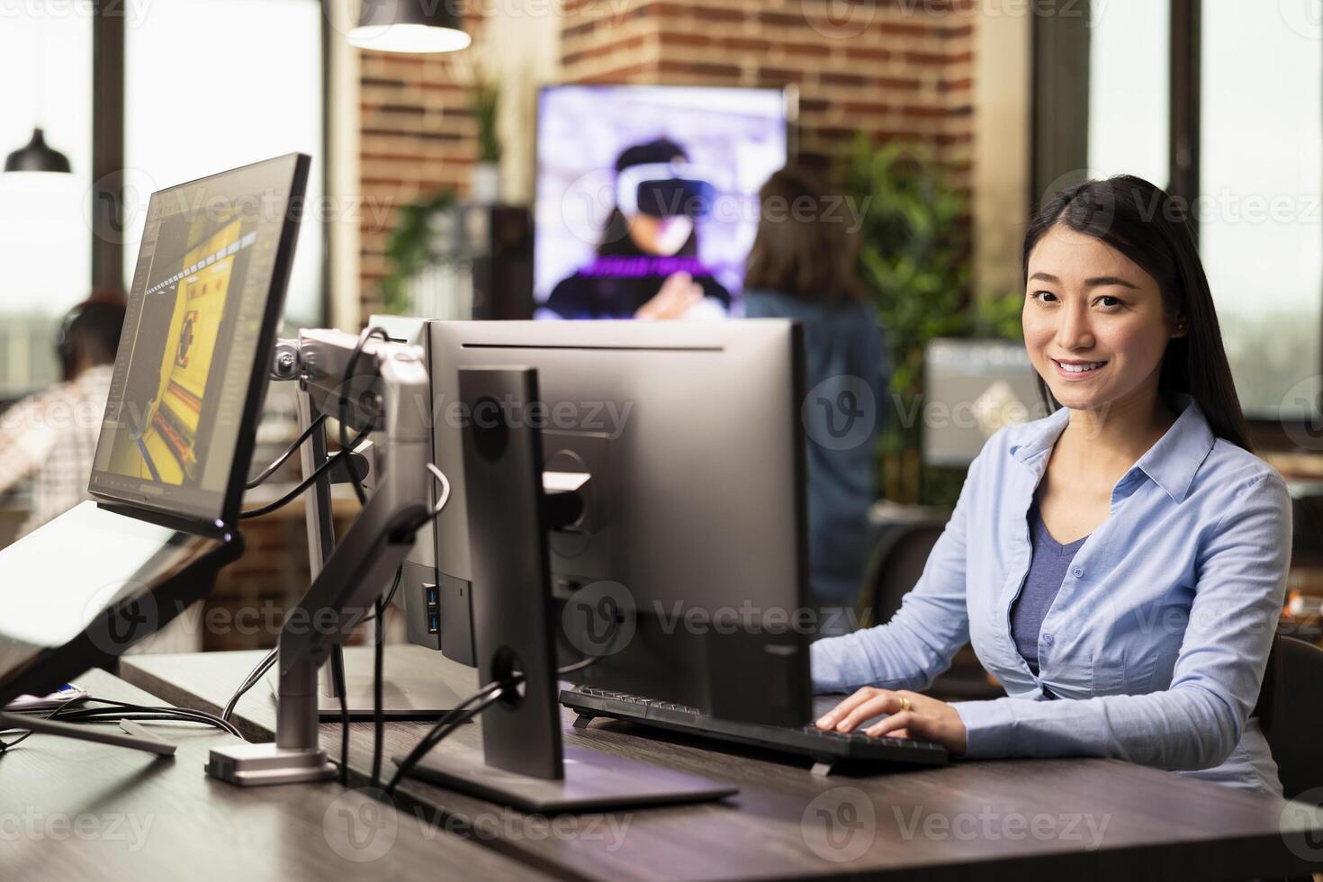 Businesswoman seated in a brick wall workspace, using desktop pc, replying to emails. Asian female creative working at her office desk, typing on a computer keyboard and smiling at the camera. photo