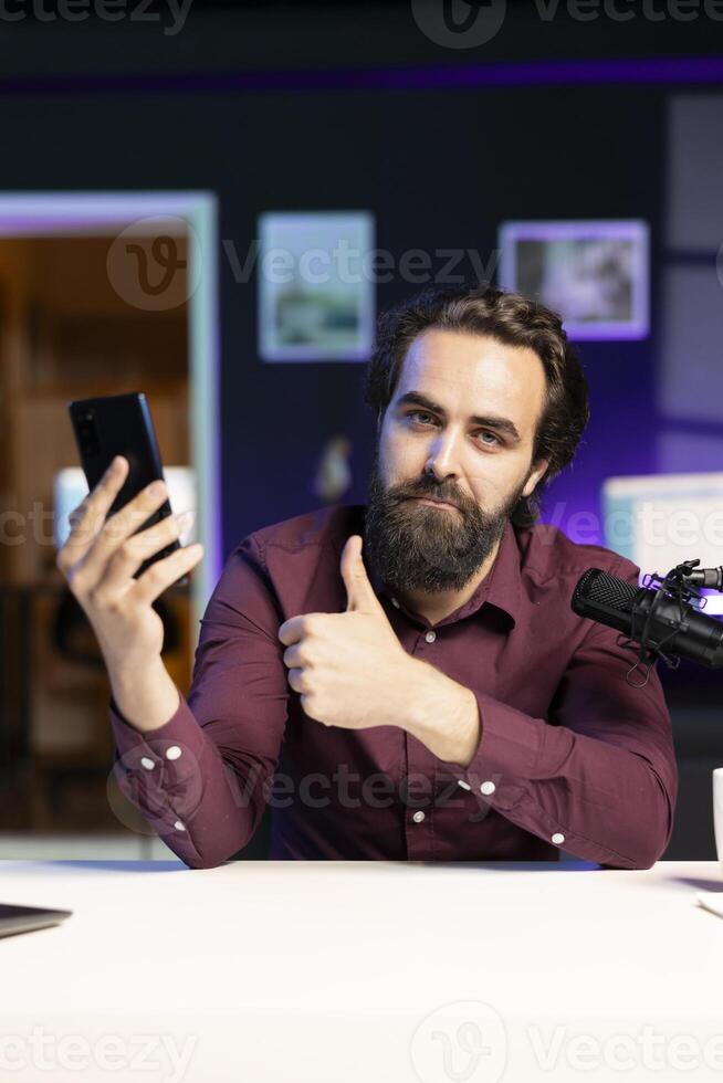 Tech guru reviewing latest phone, giving it thumbs up gesturing while filming internet show in rgb lights studio. Influencer endorsing best smartphone, happy with performance photo