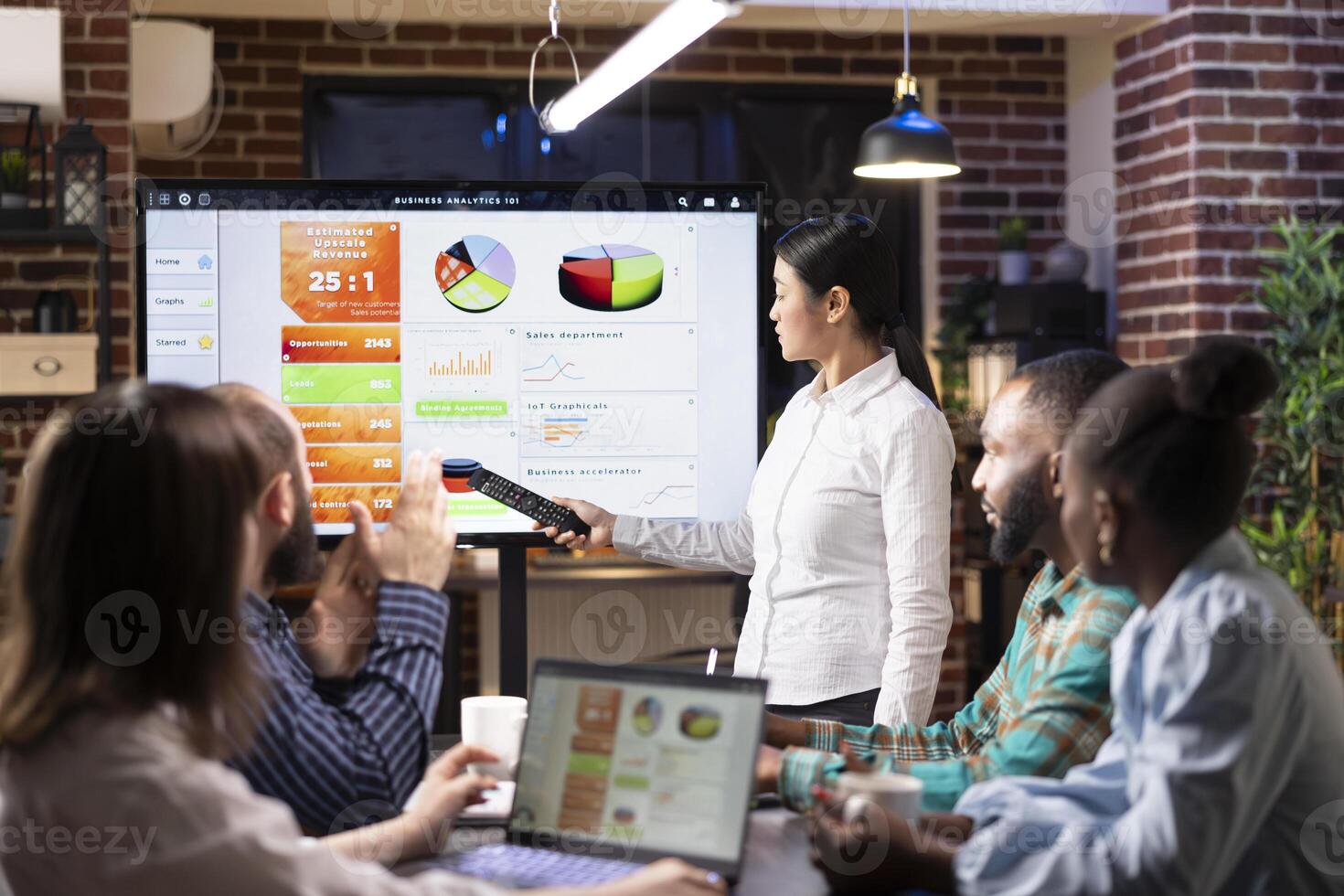 Asian businesswoman with remote in hand, pointing to screen, explaining financial data during meeting with diverse employees. Focused professionals discuss strategy using laptops and digital devices. photo
