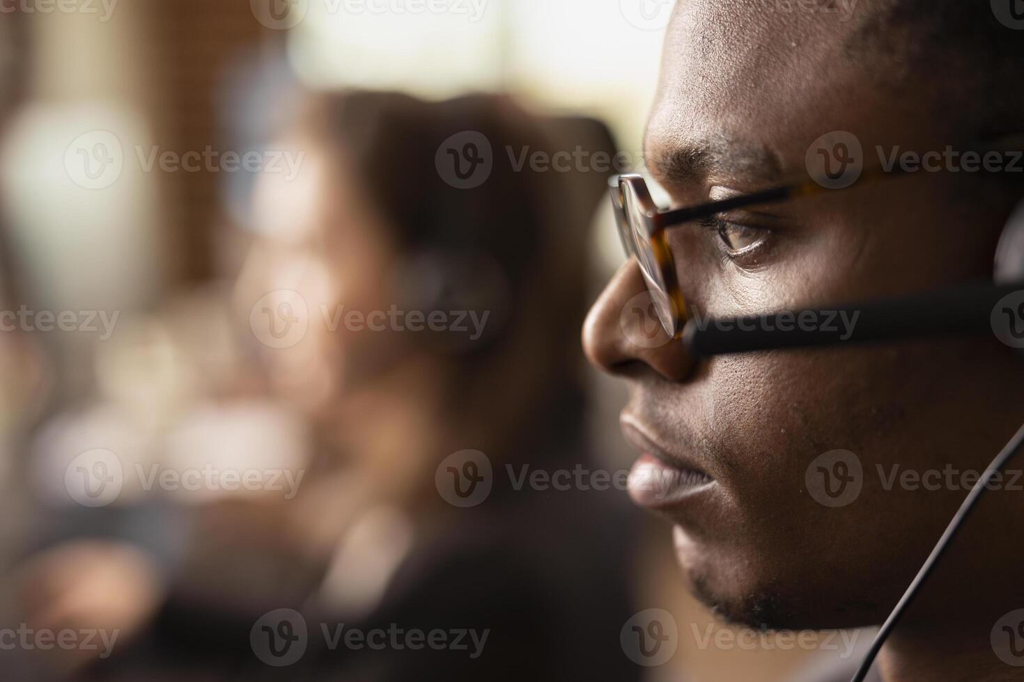 Closeup of african american man with glasses, wearing a headset with microphone while speaking with online customers. Focused male telemarketer working in startup office, providing technical support. photo
