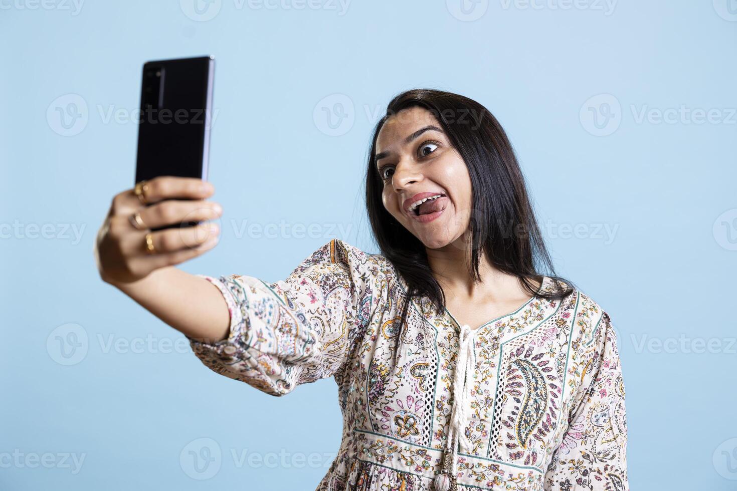 Indian woman smiling and posing for selfies on her smartphone, showcasing a positive attitude and happiness. Joyous silly person uses phone for pictures on social media, studio background. photo