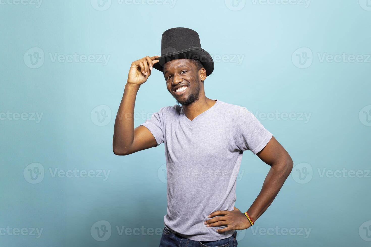 Young black man adjusting his top hat and posing for the camera against a blue background. Happy male illusionist wearing a vintage magician topper and standing with his hand on his waist. photo