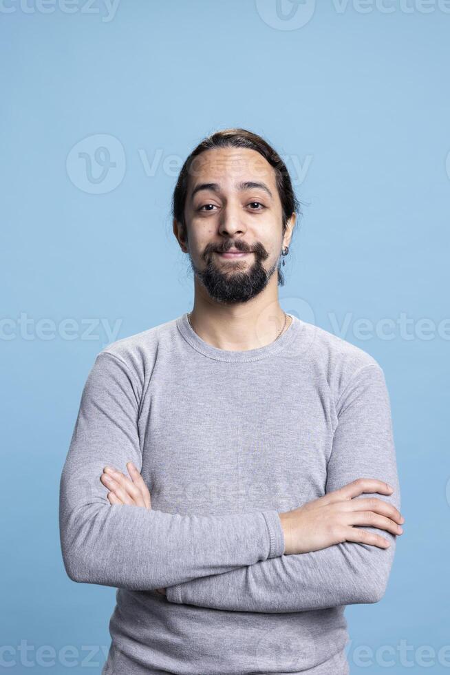 Young adult in casual wear standing with his arms crossed on camera, posing with confidence against a blue background. Man with a beard and long hair, calm natural attitude. photo