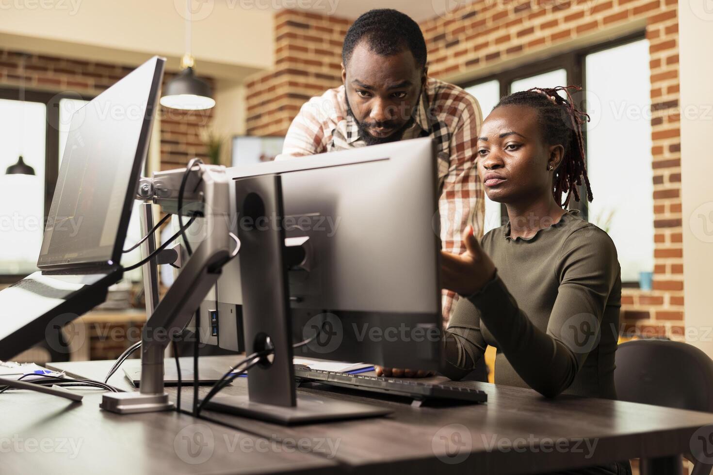 African American coworkers collaborate on product designs, exchanging ideas in brick wall office. Black man standing at desk and having creative conversation with female employee working on computer. photo