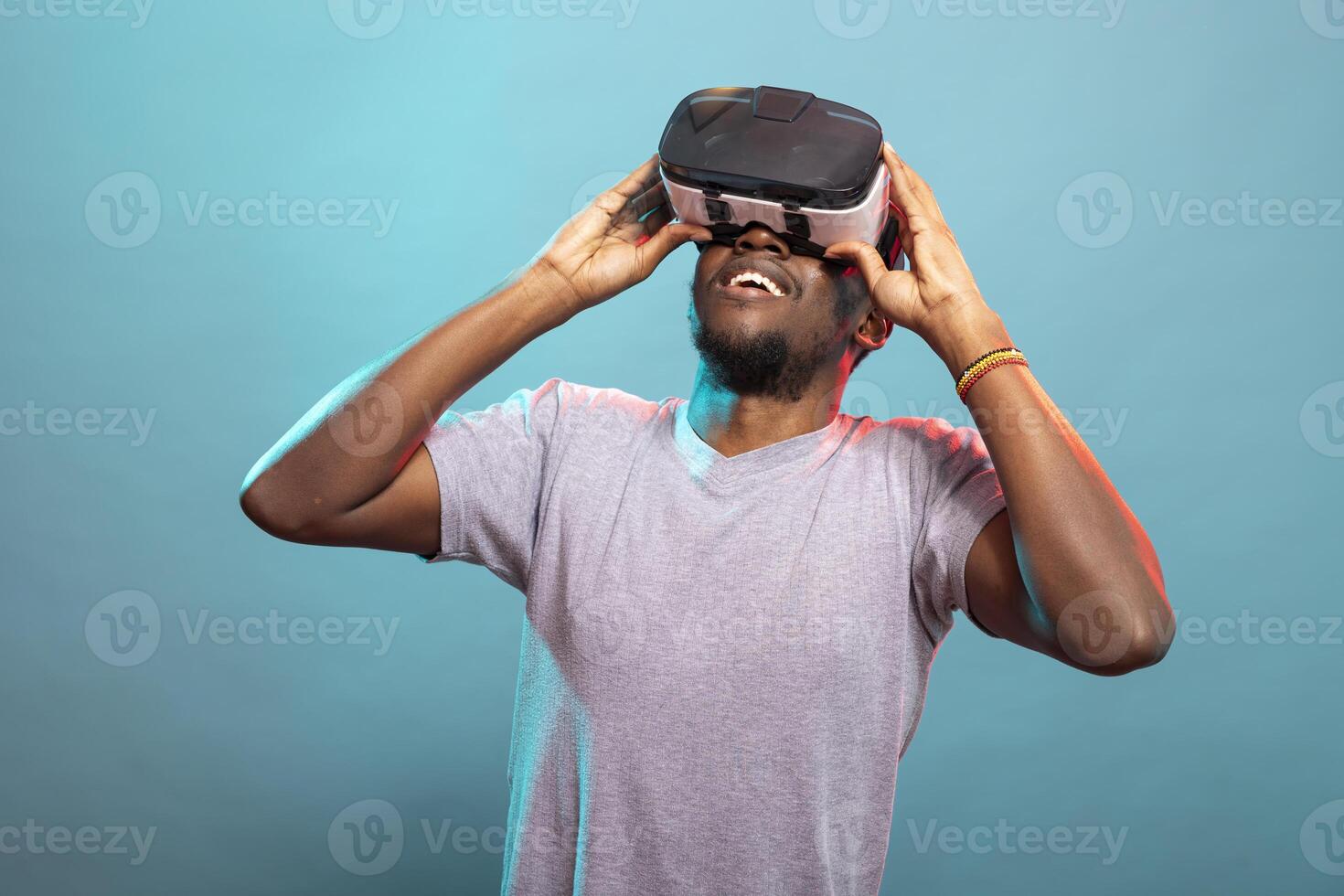 Modern adult playing an interactive 3D game against a blue background, fully immersed in virtual entertainment. Black male model touching his vr headset, looking up, exploring a futuristic simulation. photo