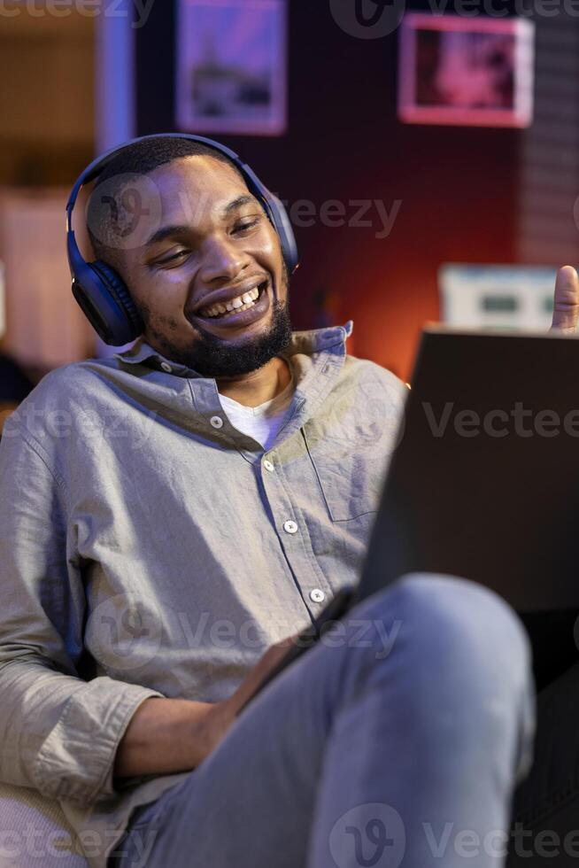 Confident male person attends a webinar lesson online on a call, paying attention and engaging in discussion remotely. Young black guy meeting with attendees on laptop, relaxing on sofa. photo