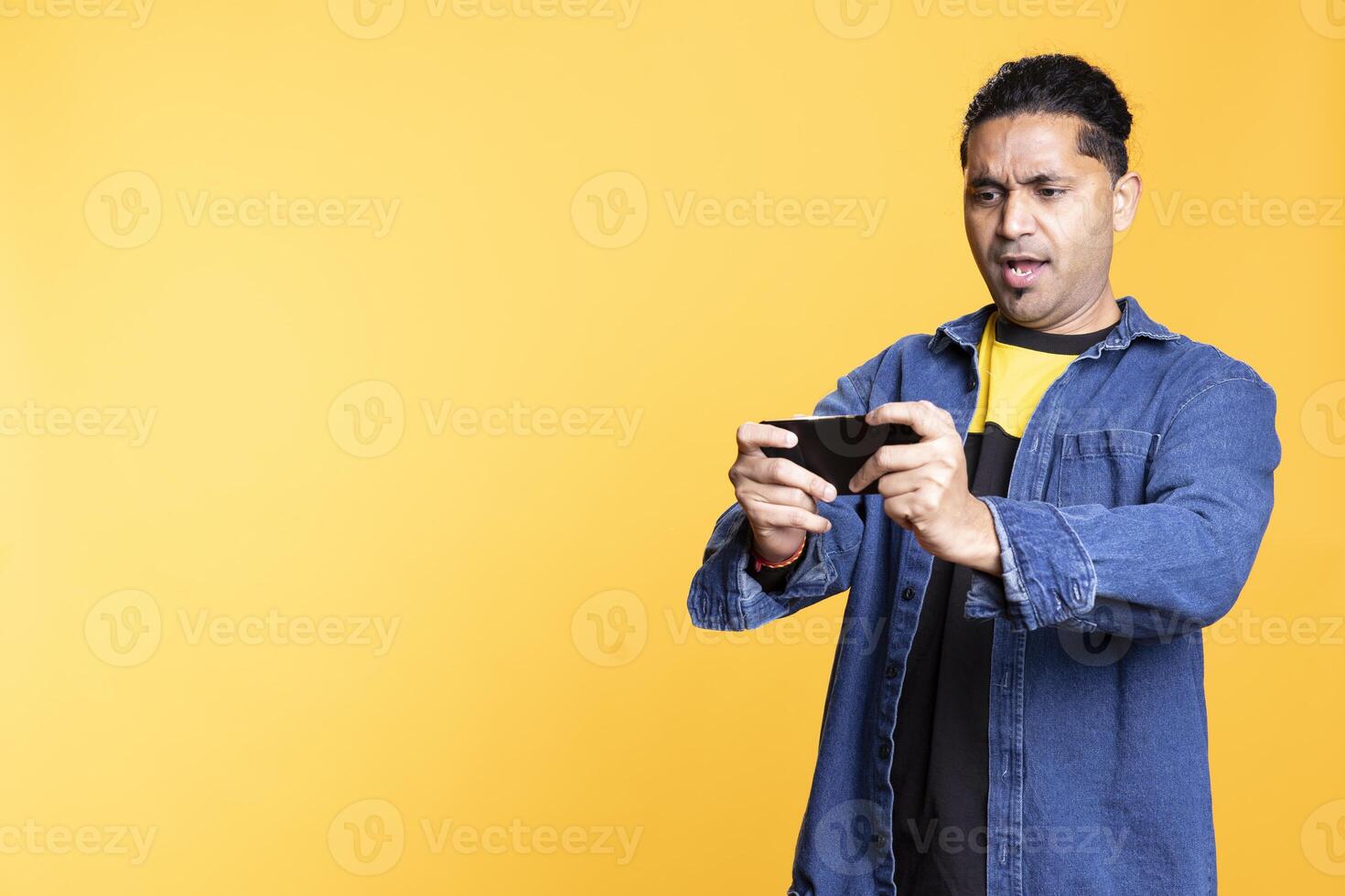 Cheerful indian person playing games on his mobile phone, celebrating after defeating foes against orange background. Young player with man bun, thrilled about online games. photo
