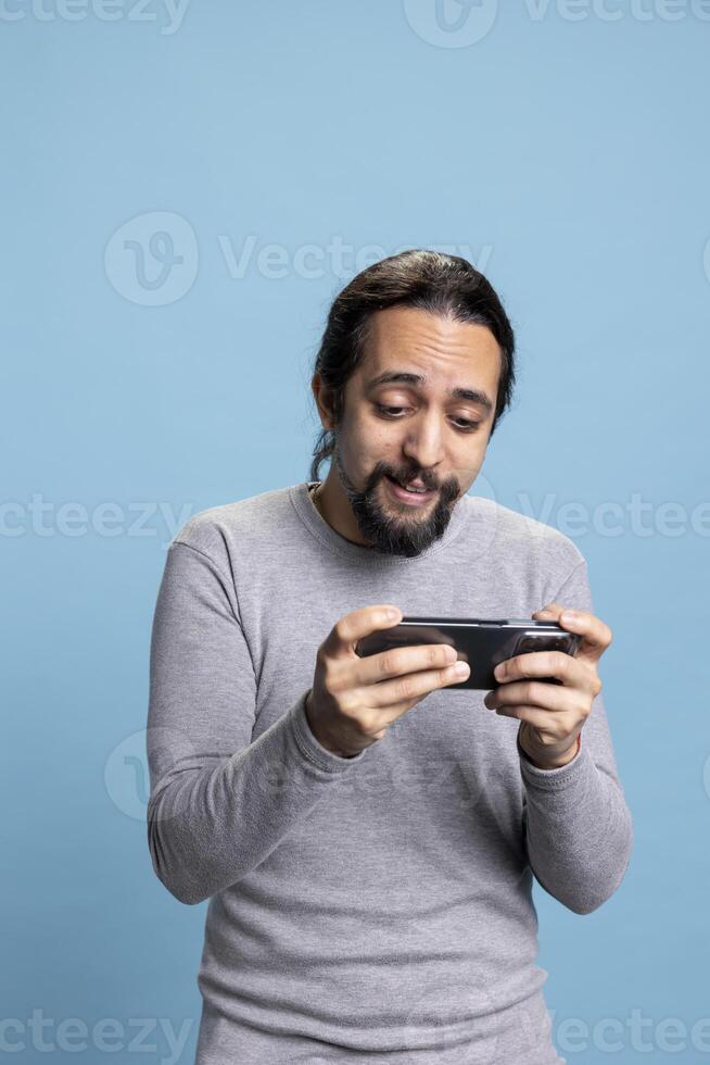 Casual modern guy playing games tournaments on mobile phone, participating in an electronic competition of a shooter game against blue background. Young person with long hair and beard. photo