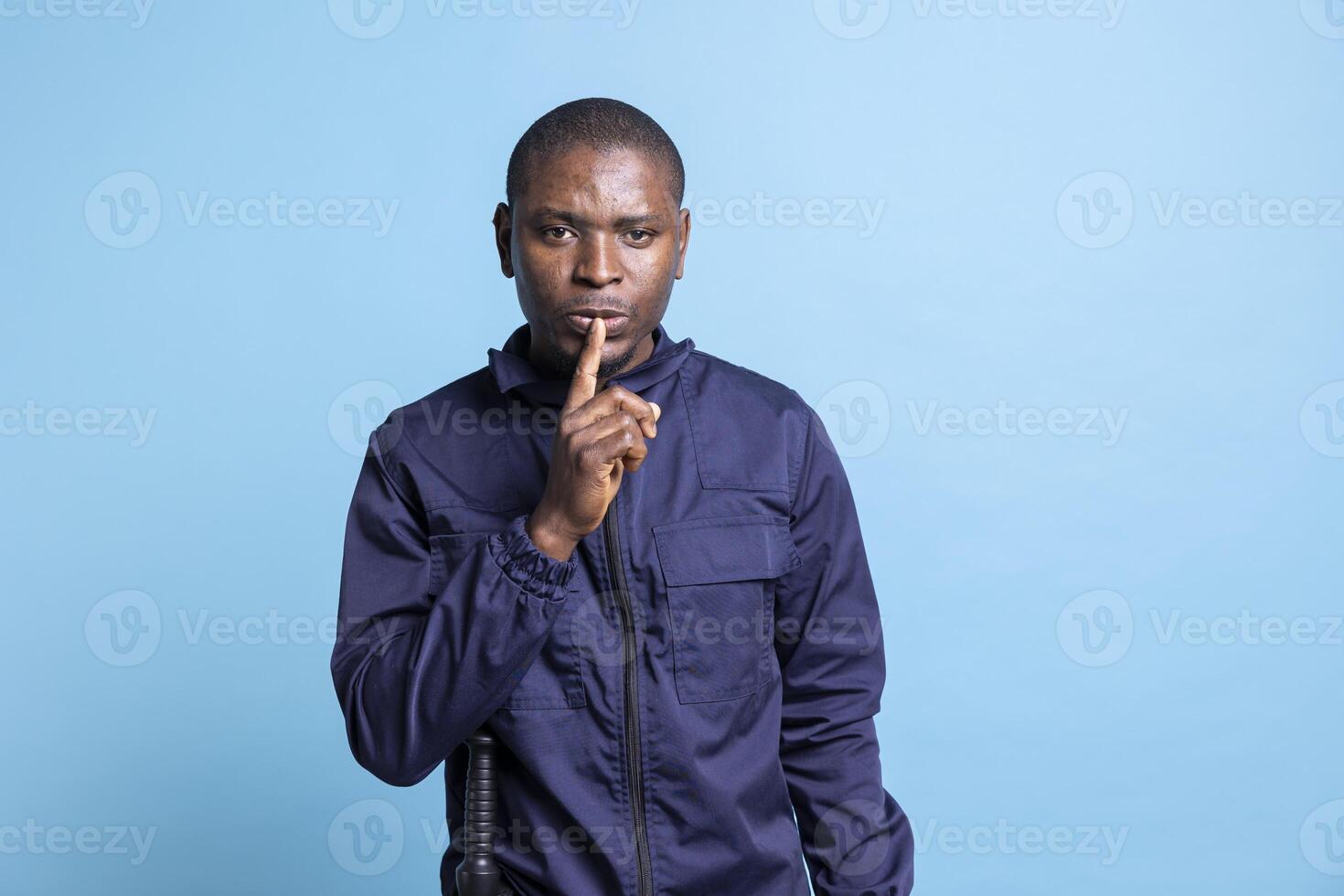 African american patrol officer putting a finger over lips for silence, presenting a hush symbol against blue background. Man in security guard uniform shows mute sign for a quiet second. photo