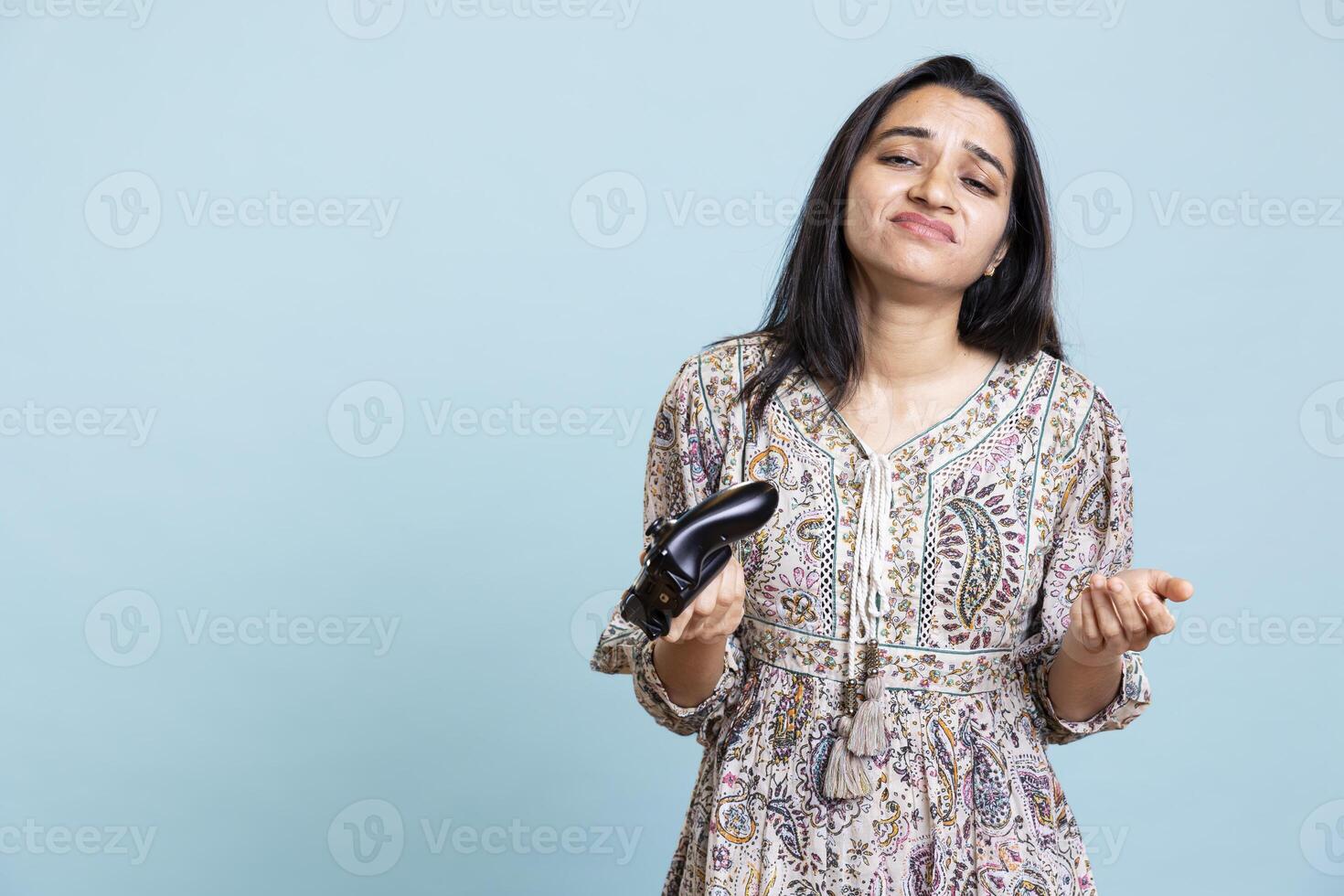 Indian girl player disappointed after losing game on console, standing against blue background. Displeased irritated woman due to lost gaming tournament, playing with controller. photo