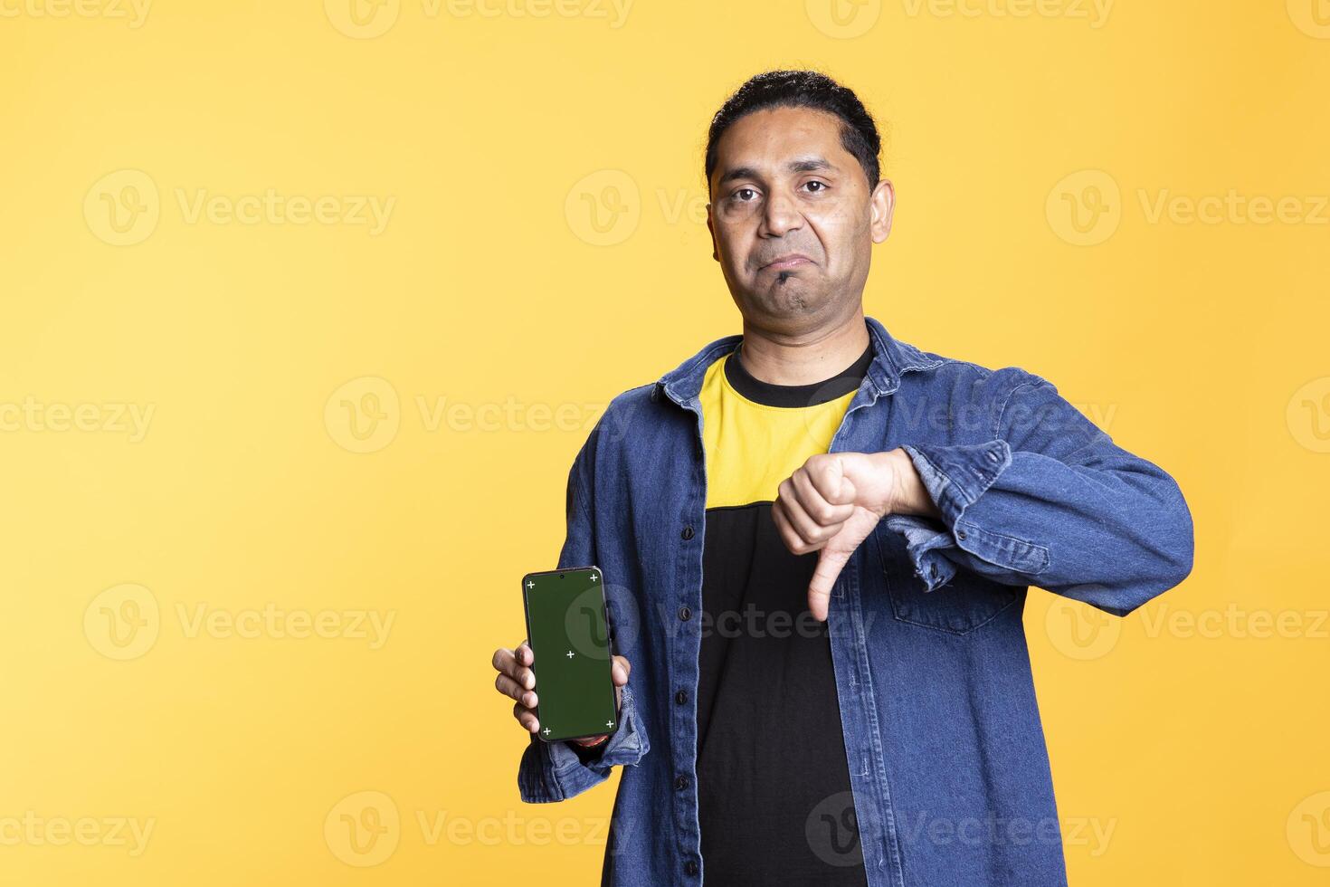 Young adult shows thumbs down next to copy space on his phone, creating an advertisement for marketing against orange background. Indian person in a positive mood, green screen. photo