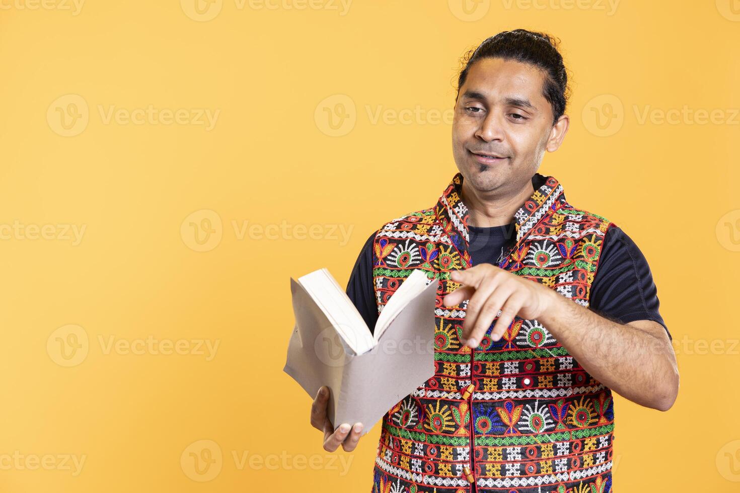 Jolly indian teacher reading book for pupils during class, pointing finger gesture, studio background. Happy man holding novel, telling story to kindergarten kids, asking them questions photo