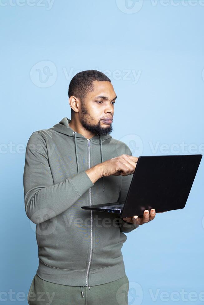 Carefree happy african american guy working on his laptop in studio, wearing a grey hoodie and composing emails to solve his tasks and paperwork. Young adult multitasking, browses websites. photo
