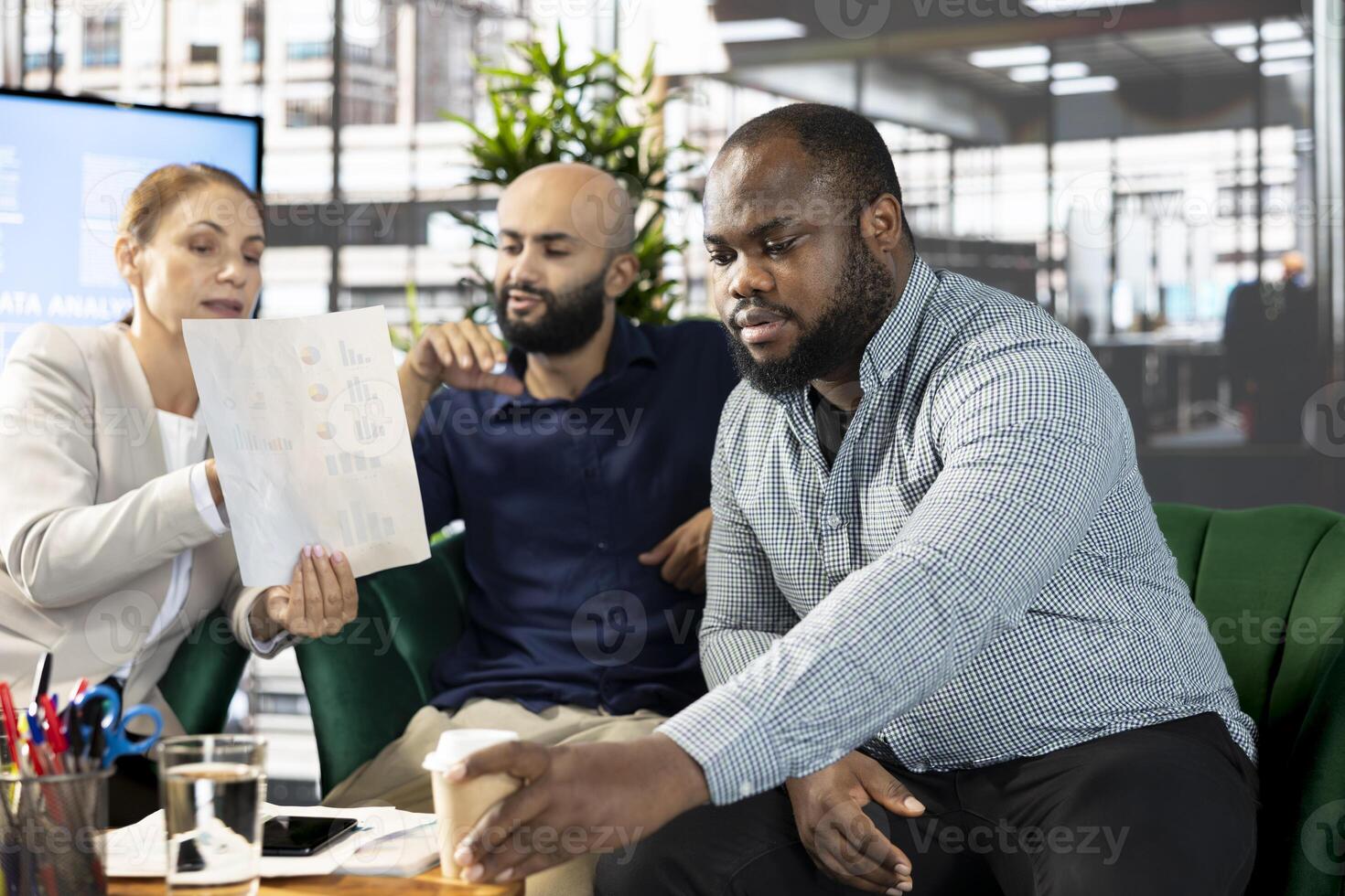 Man drinking coffee next to team of office clerks looking over revenue figures on documents. African american employee enjoying beverage next to staff members working hard photo