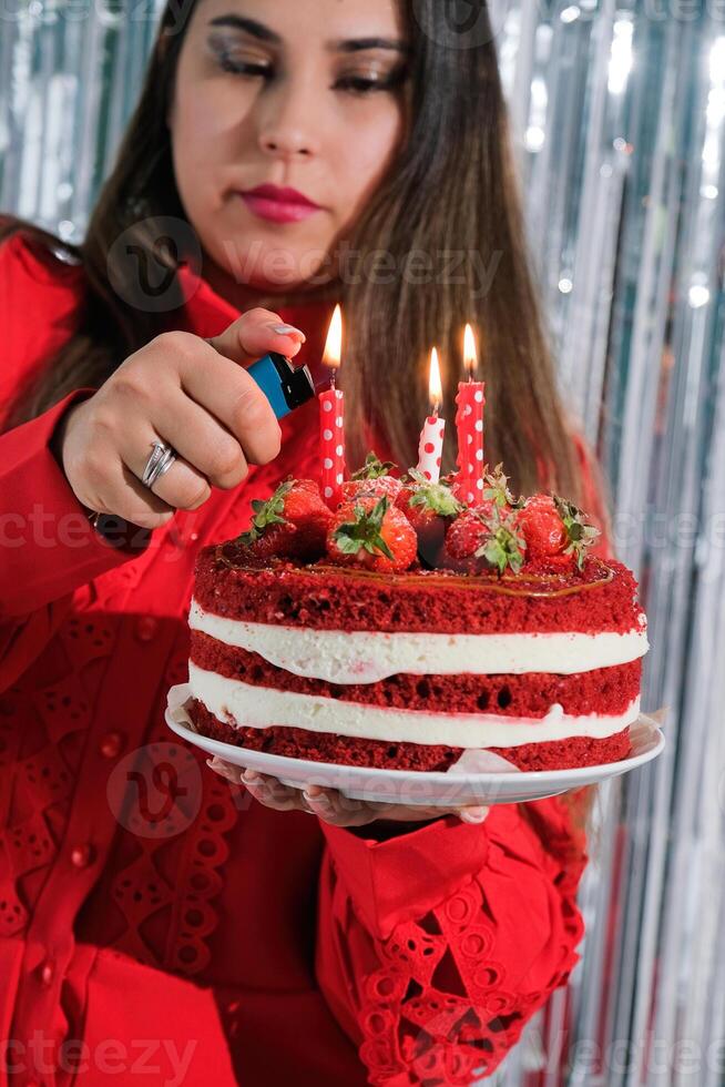eautiful woman wearing red dress celebrating birthday and lighting candles on red velvet cake, making a wish photo