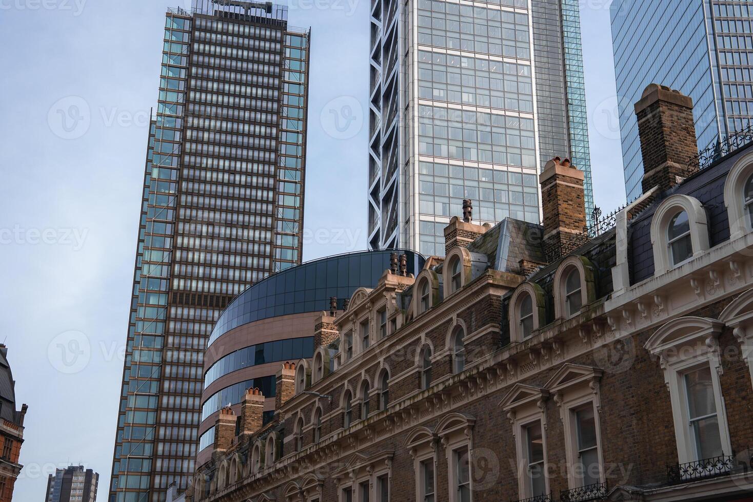 A traditional brick building with chimneys stands in contrast to sleek skyscrapers, including a cylindrical and grid patterned tower, under a clear sky in London. photo