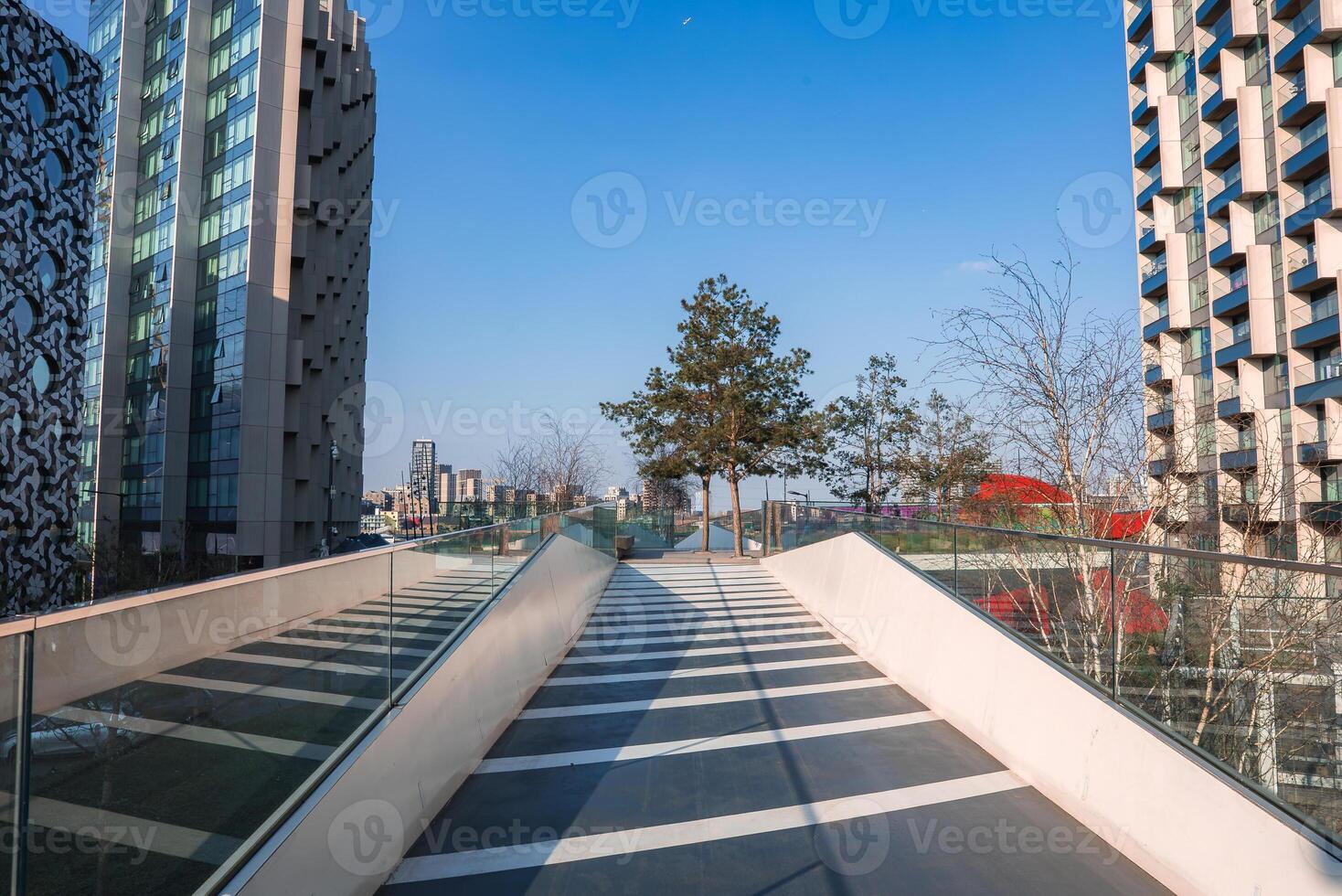 A glass railed walkway leads to trees and an open area, surrounded by high rise buildings with unique facades under a clear blue sky in London. photo