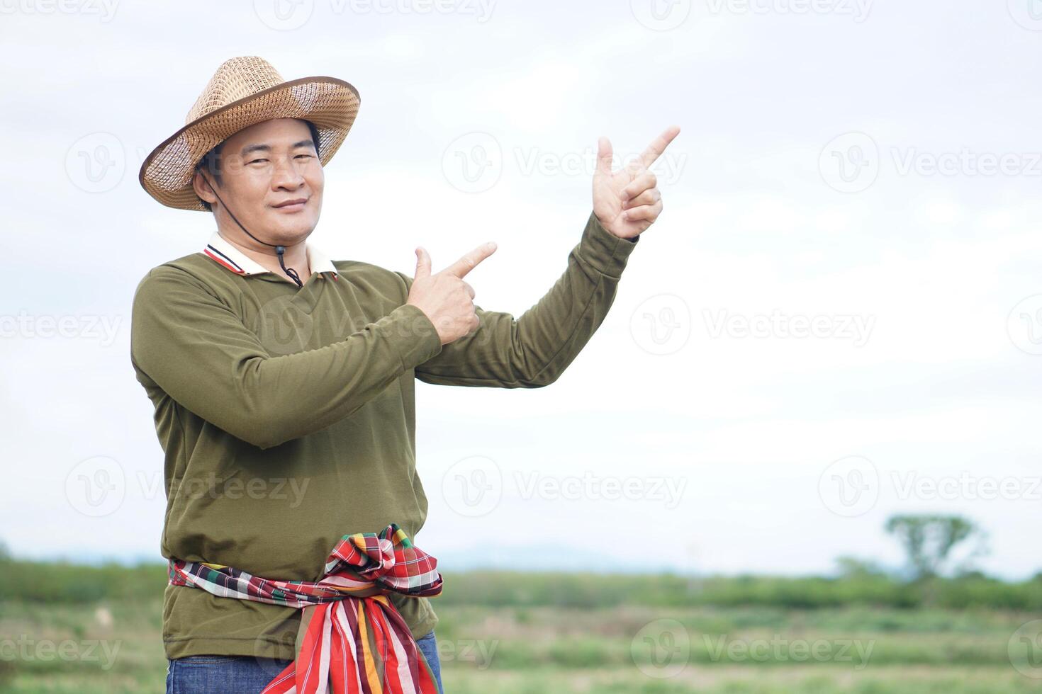 Handsome Asian man farmer stands at agricultural land, wears hat, Thai loincloth on waist, point finger up, feels confident. Concept agriculture occupation. photo