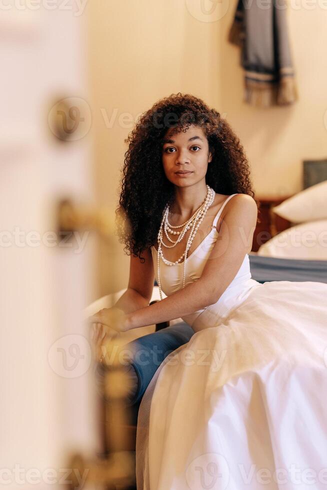 Portrait of a cute African woman in a white elegant dress and jewelry. Attractive curly black girl sitting on a retro sofa. Charming teenager relaxing in a hotel room. photo