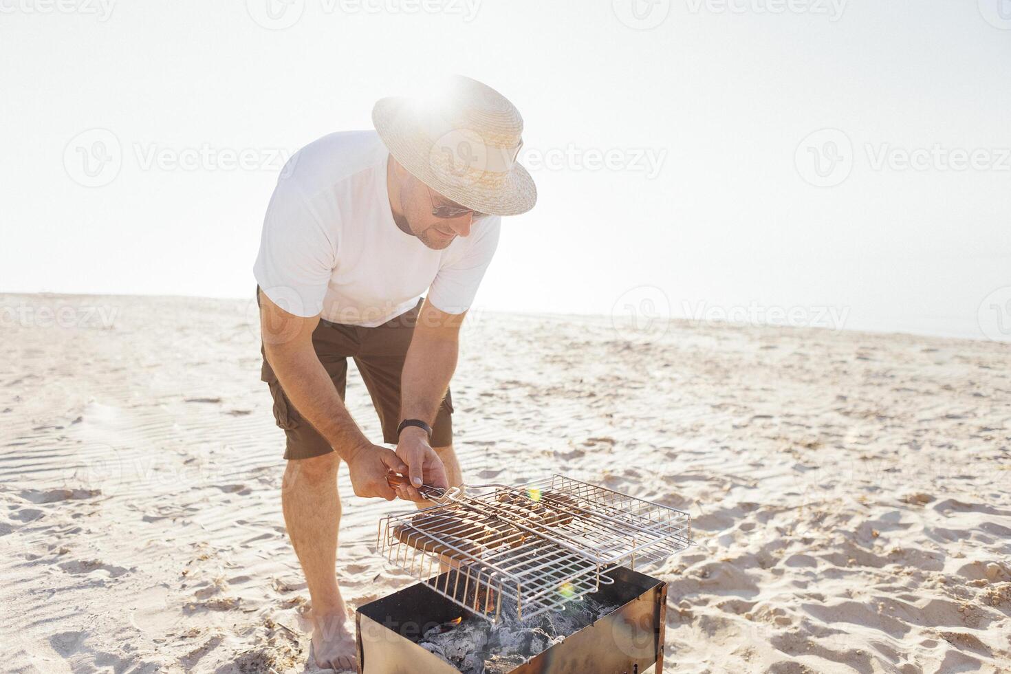 A young man in a summer casual outfit is having a barbecue on the sandy beach. A male in sunglasses and a straw hat is frying sausages on the seashore. photo
