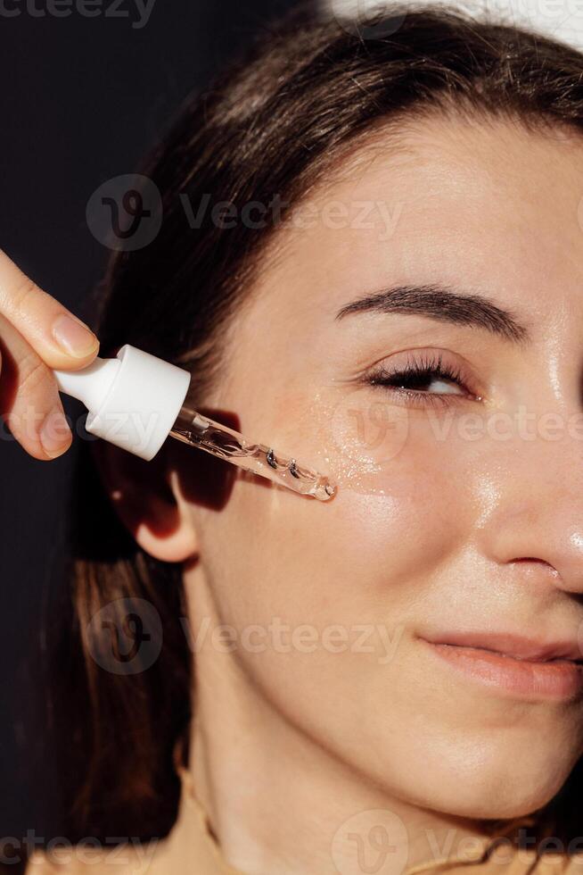 Close-up portrait of a young girl applying a transparent essence to her face. A teenager holds a dropper with a moisturizing serum. Skin care. Bright sunlight. photo