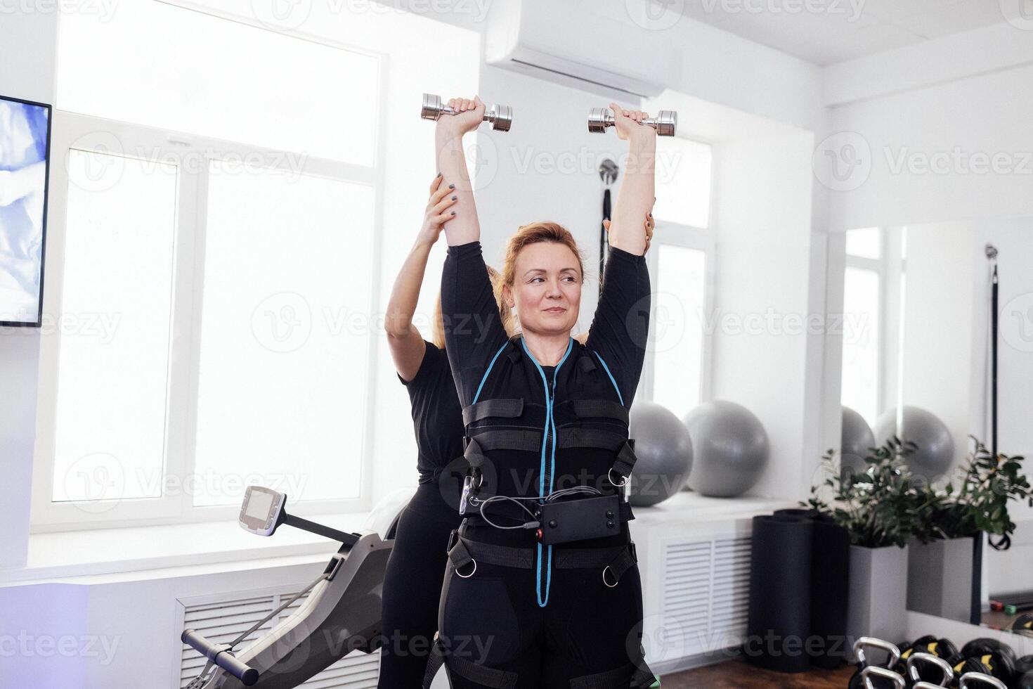 Smiling middle age sportswoman in ems suit is engaged in physical activity with personal trainer. photo