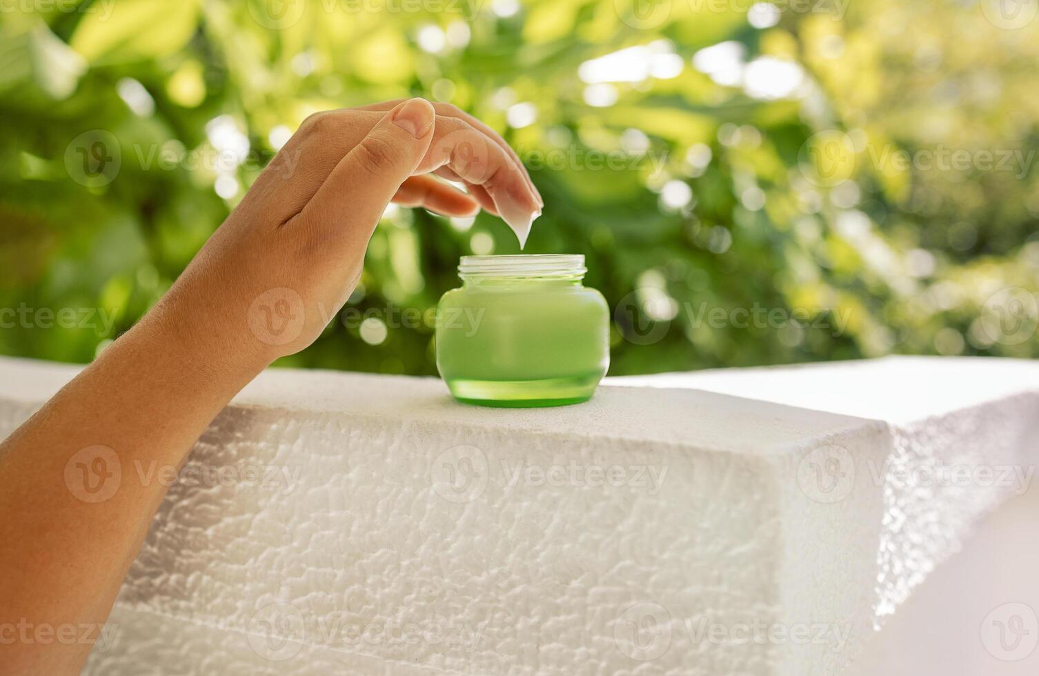 Close-up of a woman hand with white moisturizer on her fingers and a green jar. Cosmetic product with anti-aging cream on the background of bright green foliage. photo