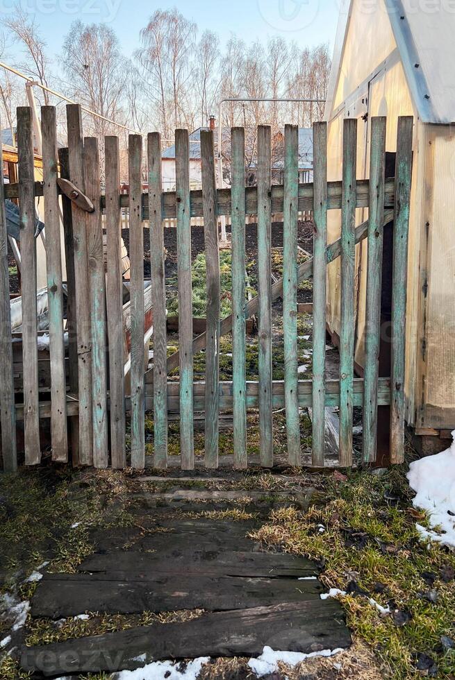 Rustic wooden garden gate leading to a backyard area during early spring. The scene captures remnants of snow melting on the ground, with bare trees in the background photo