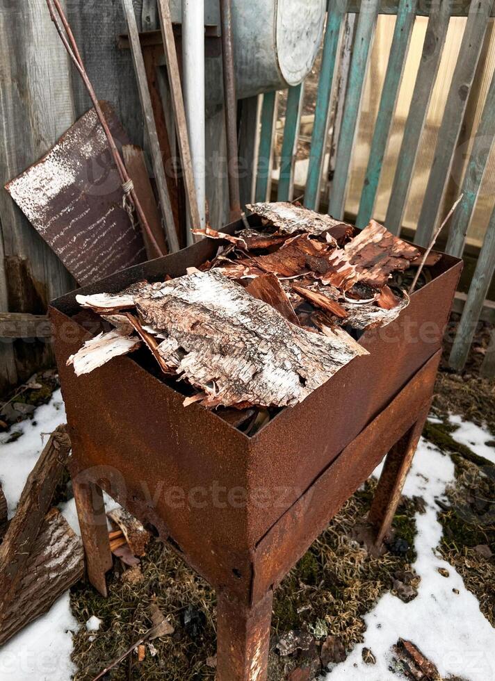 Rusty metal grill filled with dry birch bark, ready for lighting a barbecue fire. The setup is outdoors, surrounded by rustic materials photo