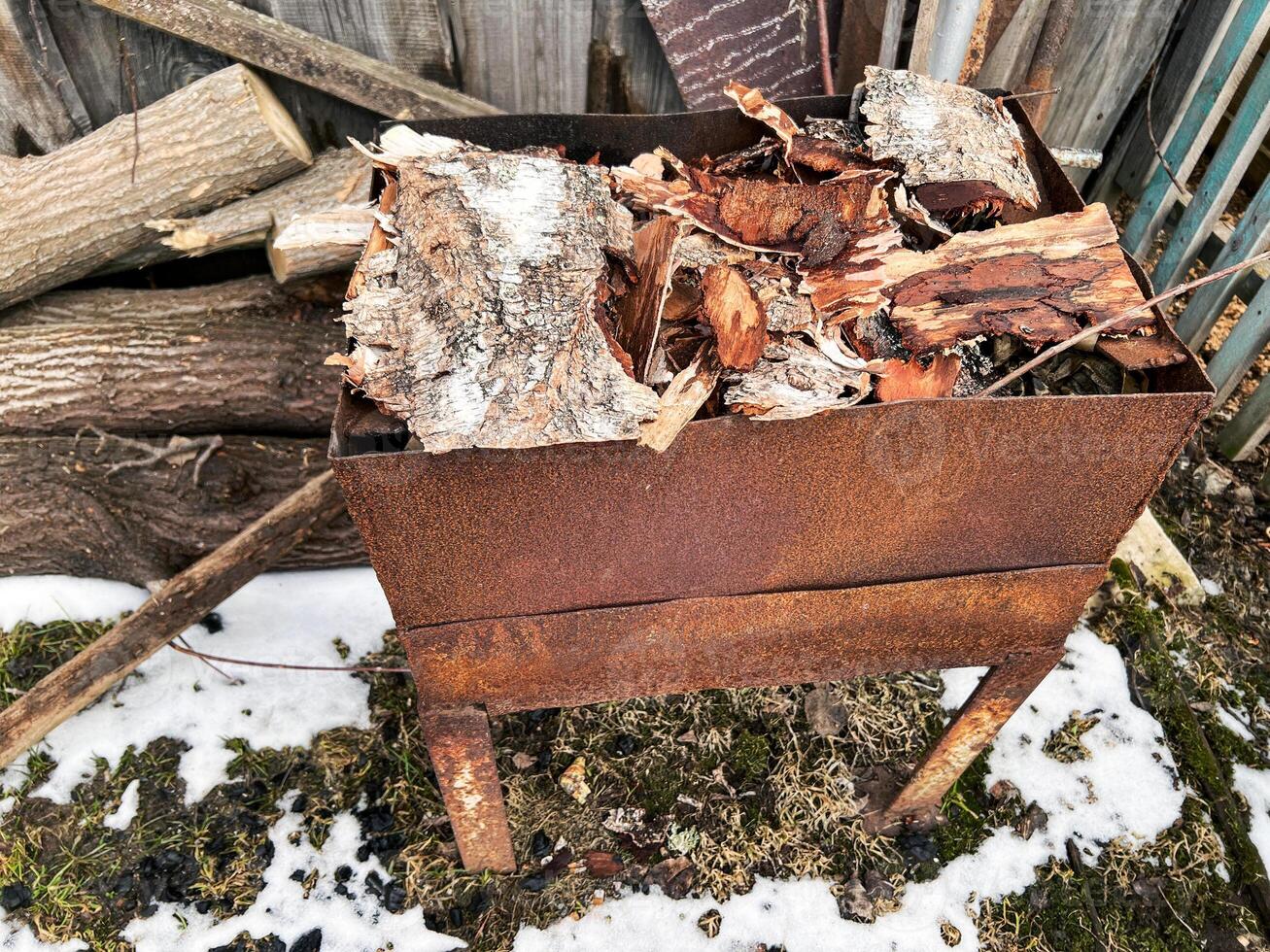 Rusted metal fire pit filled with birch logs and wood pieces. Surrounded by logs and patches of snow, the scene highlights a rustic outdoor setting, perfect for backyard fires and winter warmth photo