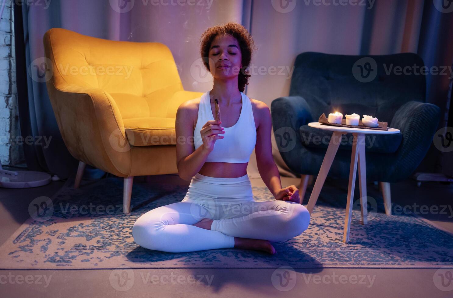 Young African in fitness outfit is sitting on floor in living room and enjoying scent of incense stick photo