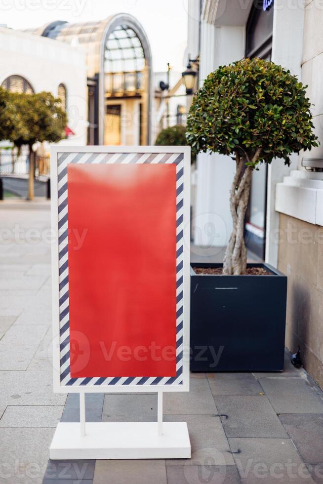 Blank advertising board stands on sidewalk near trees and modern architecture in daytime photo
