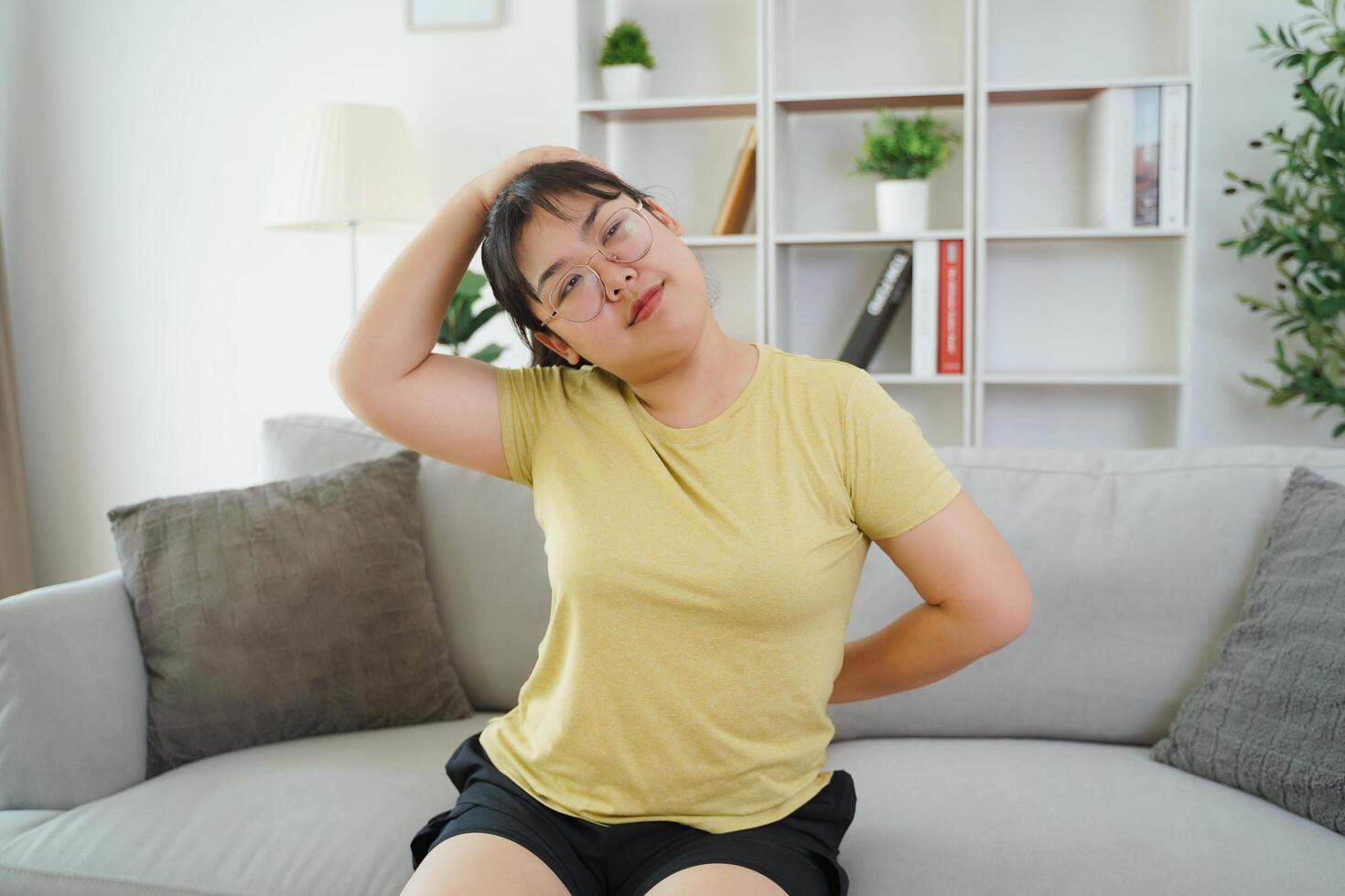 plump Asian woman stretching her muscle while sitting on a sofa in her living room at home. photo