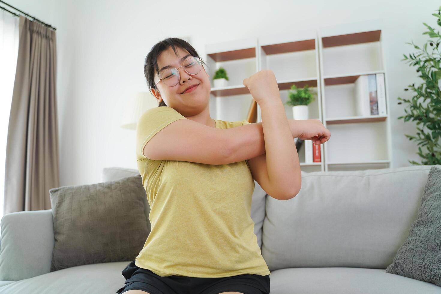 A plump woman is stretching her muscles while sitting on a sofa in her living room at home. photo