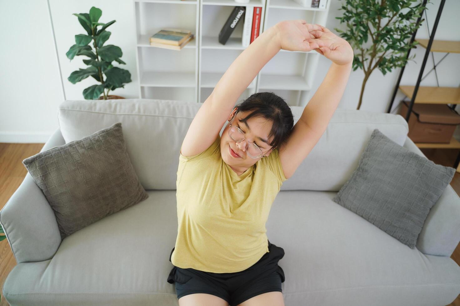 plump Asian woman stretching her muscles while sitting on a sofa in her living room at home. photo