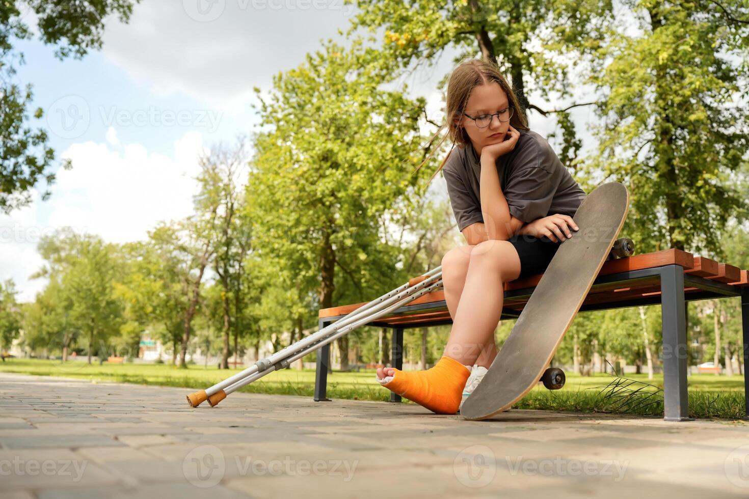 teenage girl, her leg in a cast, sits on a park bench, her gaze fixed on the skateboard that led to her injury, a bittersweet moment of reflection photo