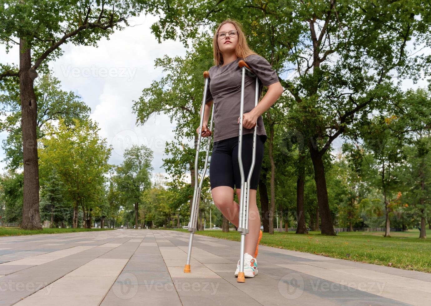 Young girl with a bright orange cast on her broken leg stands upright on a brick path in a lush summer park, utilizing crutches to carefully navigate the outdoor environment photo