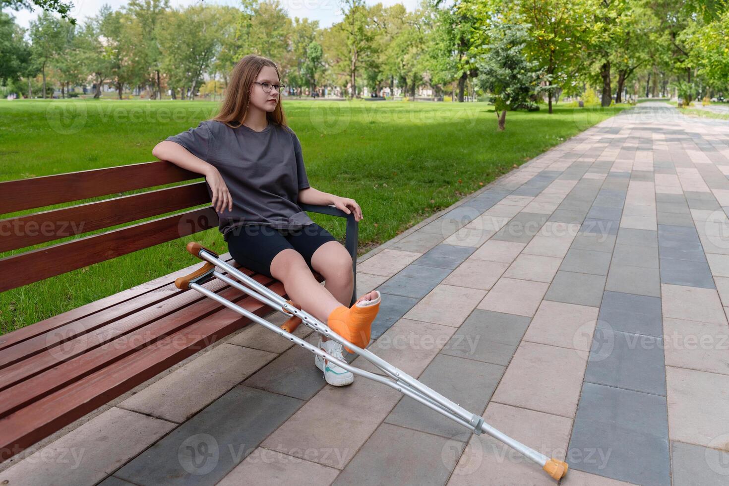 Teenage girl with a leg in a cast seated on a bench in a scenic summer park, her crutches providing mobility support during her recovery photo
